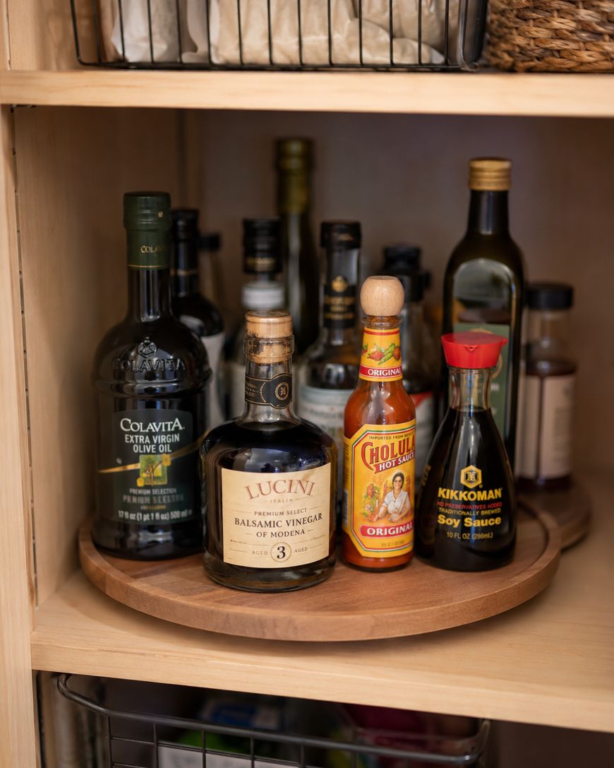 A wooden lazy Susan holds bottles of olive oil, balsamic vinegar, hot sauce, and soy sauce inside a pantry cabinet, with other condiments and bottles in the background.