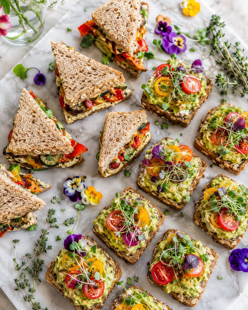 An assortment of open-faced sandwiches with avocado, cherry tomatoes, microgreens, and edible flowers, alongside stacked sandwich triangles with whole grain bread and colorful vegetable fillings, arranged on a marble surface.