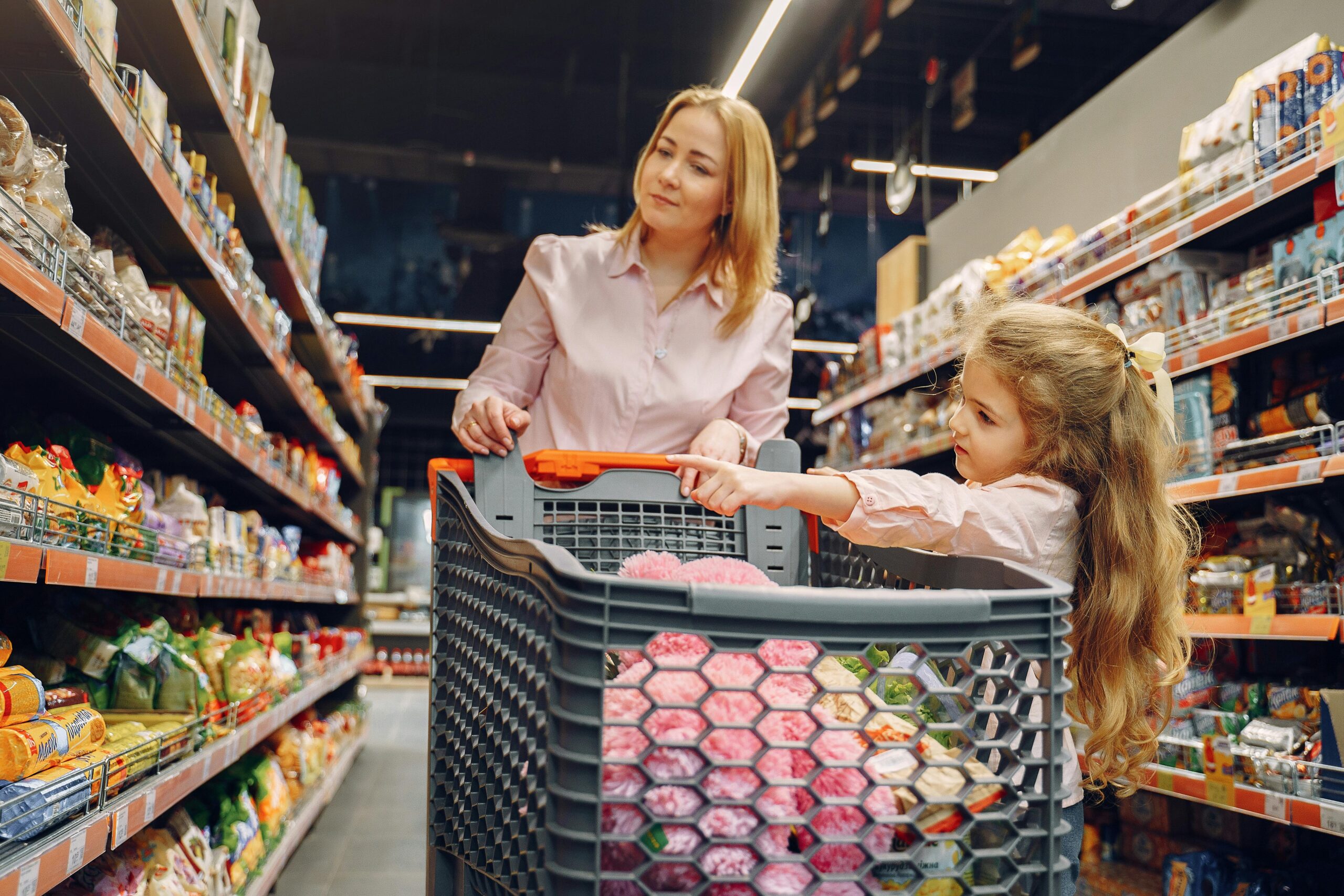A woman and a young girl shop in a grocery store aisle. The woman pushes a cart filled with pink items while the girl points at products on the shelves. Both appear to be focused on grocery shopping.