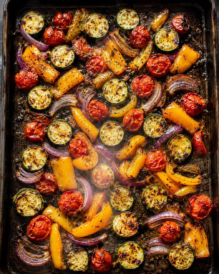 A baking tray filled with roasted vegetables including sliced zucchini, cherry tomatoes, yellow bell peppers, and red onion, all seasoned with herbs and spices.
