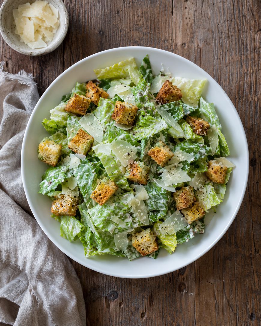 A bowl of Caesar salad with fresh romaine lettuce, golden croutons, and shaved Parmesan cheese sits on a rustic wooden table next to a small bowl of extra cheese and a beige cloth napkin.