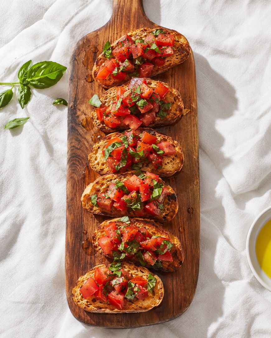 Five pieces of bruschetta topped with diced tomatoes and fresh basil are arranged on a wooden serving board, with a sprig of basil and a bowl of olive oil nearby.