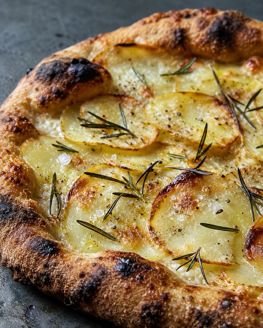 Close-up of a golden-brown potato pizza topped with thin potato slices, rosemary, black pepper, and sea salt on a crispy, charred crust.