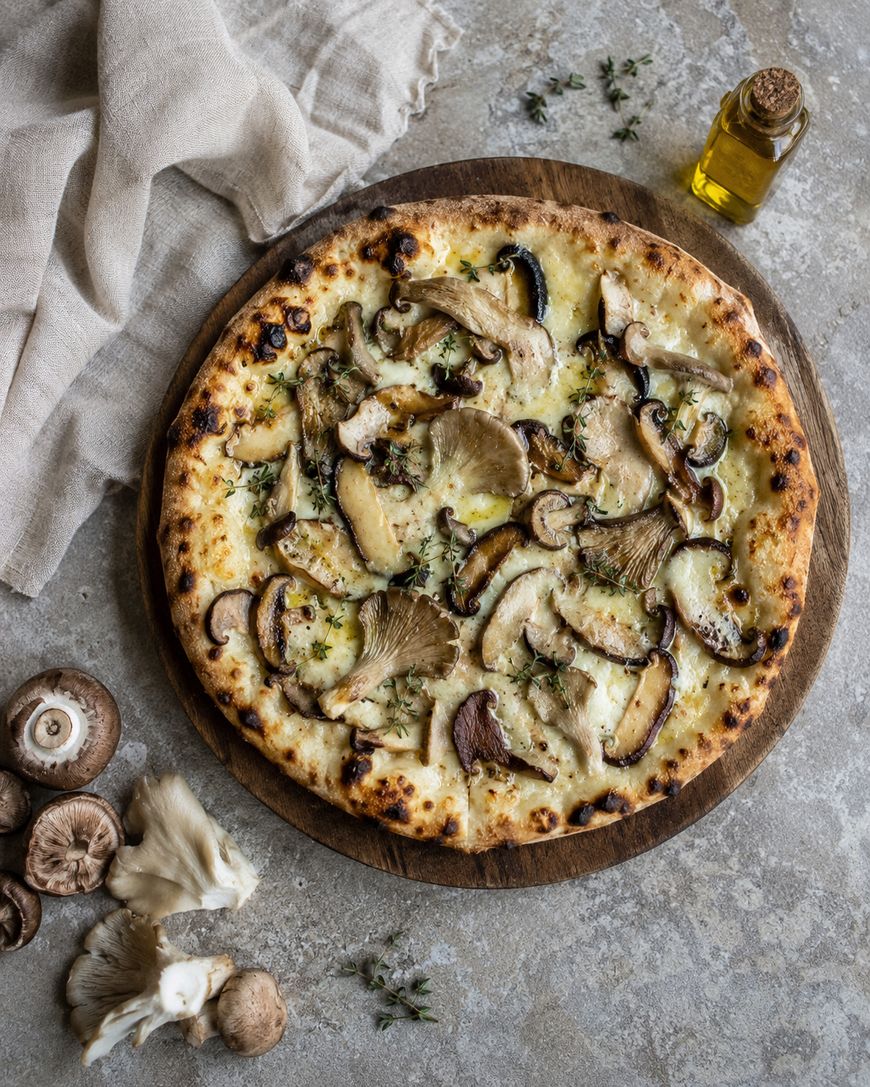 A wood-fired pizza topped with various mushrooms, melted cheese, and herbs sits on a round wooden board. Fresh mushrooms, thyme, a linen cloth, and a small bottle of olive oil are nearby on a textured surface.
