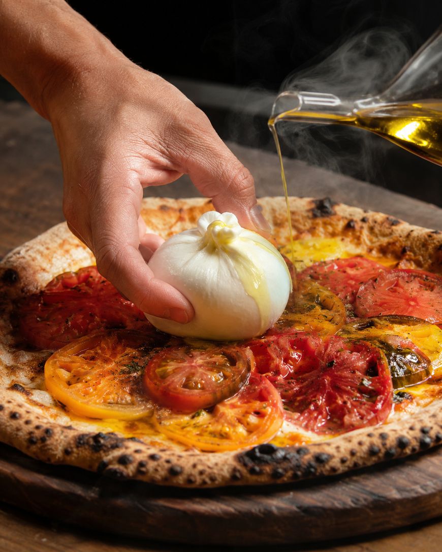 A hand places a ball of burrata cheese on a tomato pizza while olive oil is being poured over the top; steam rises from the freshly baked pizza on a wooden surface.