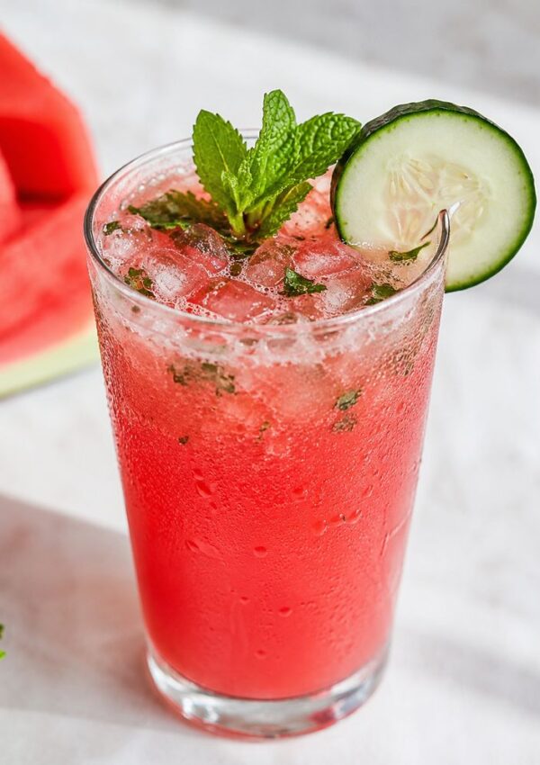 A tall glass of bright pink watermelon drink with crushed ice, garnished with a sprig of mint and a cucumber slice—perfect for crowd cocktails. In the background, a blurred piece of watermelon rests on a white surface.