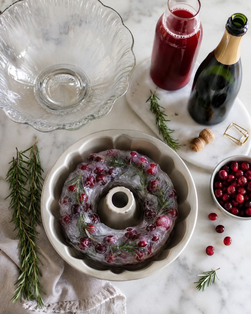 A ring-shaped ice mold with cranberries and rosemary sits in a bundt pan on a marble countertop, surrounded by a punch bowl, rosemary sprigs, a bottle of champagne, a glass bottle of red juice, and a bowl of cranberries.