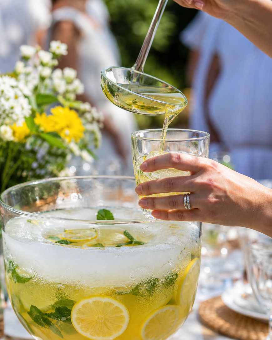 A person ladles refreshing lemonade with lemon slices and mint from a large glass bowl into a glass. Flowers and blurred people in white clothing are seen in the background at an outdoor gathering.
