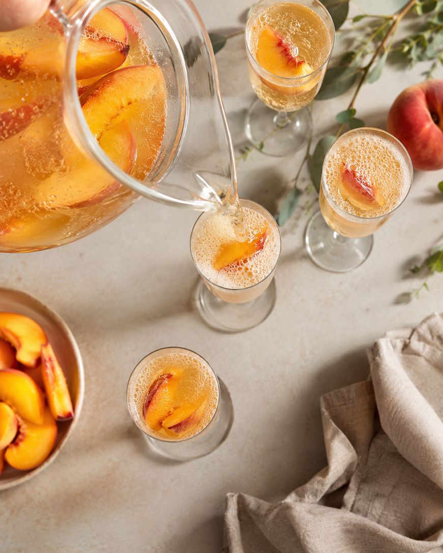 A pitcher of sparkling peach drink is being poured into a glass, surrounded by three other glasses, all garnished with peach slices. A bowl of peach slices, a peach, and a light cloth are also on the beige table.