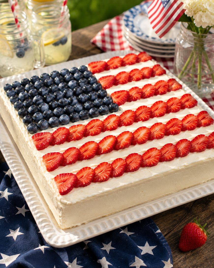 A square cake decorated like the American flag with blueberries for the blue field and sliced strawberries for the red stripes, on a white platter with a star-patterned cloth beneath.