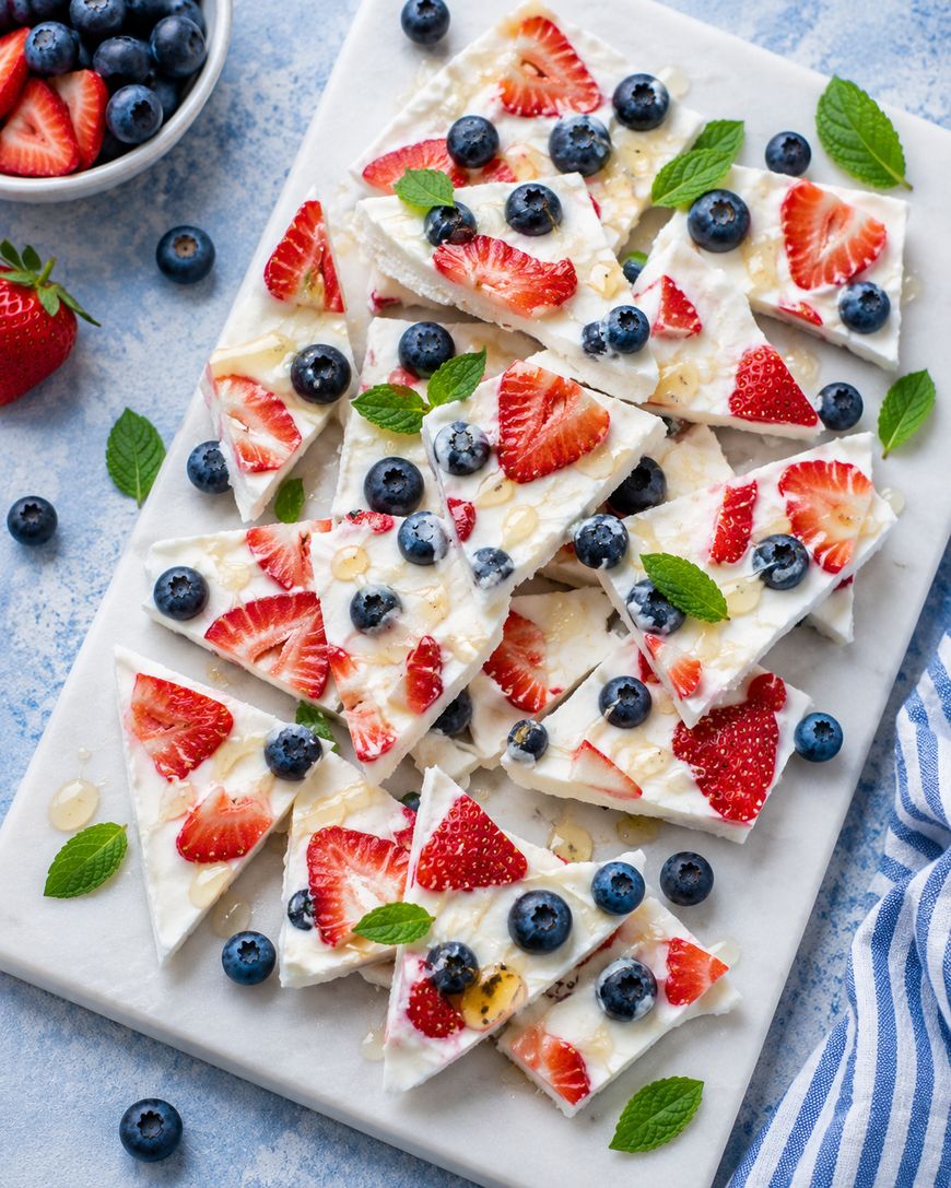 Rectangular pieces of yogurt bark topped with fresh blueberries, strawberry slices, and mint leaves are arranged on a white marble slab. A small bowl of blueberries and a striped cloth are nearby on a blue surface.