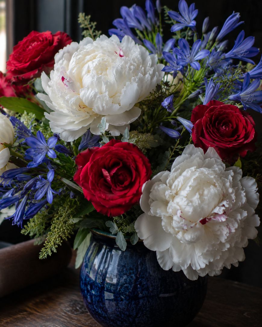 A dark blue ceramic vase holds a bouquet of white peonies, red roses, and purple-blue flowers, set against a dark background with some natural light coming from the side.