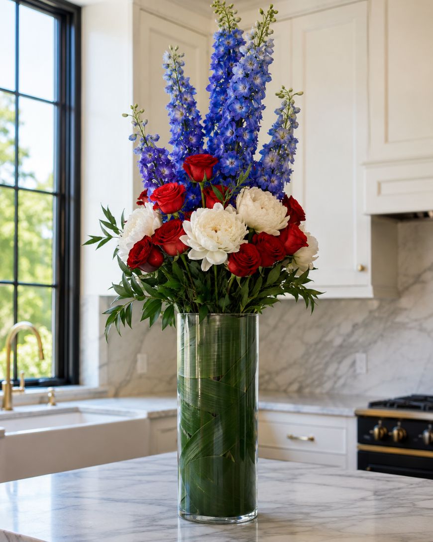 A tall glass vase filled with red roses, white peonies, and tall blue delphiniums stands on a marble kitchen island, with white cabinets and a large window in the background.