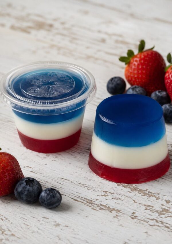 Layered red, white, and blue gelatin dessert with fresh strawberries and blueberries on a white wooden surface. One is in a clear plastic cup with a lid, the other unmolded—perfect for serving as festive Red White and Blue Jello Shots.