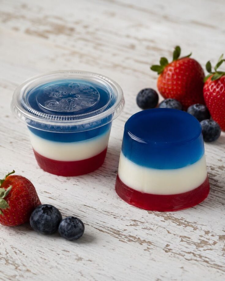 Layered red, white, and blue gelatin dessert with fresh strawberries and blueberries on a white wooden surface. One is in a clear plastic cup with a lid, the other unmolded—perfect for serving as festive Red White and Blue Jello Shots.