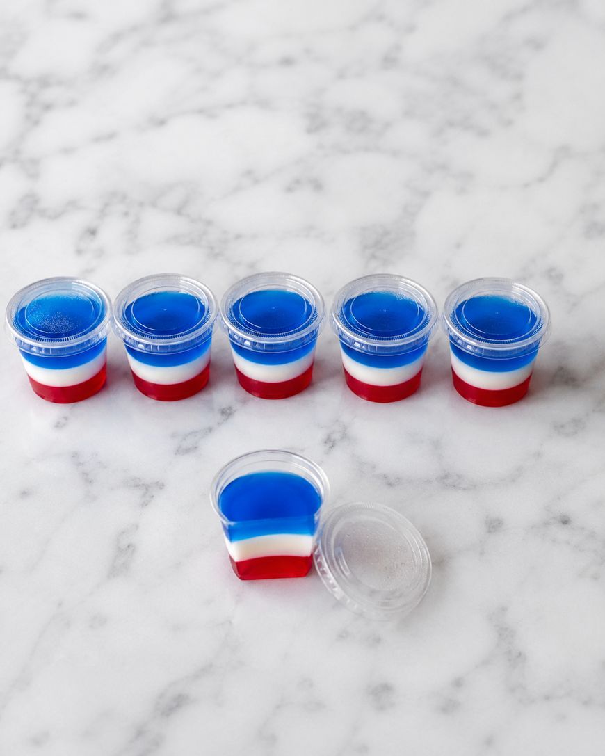 Six small plastic cups with lids are filled with layered red, white, and blue gelatin, arranged in a row on a white marble surface. One cup is open, showing the colorful layers.