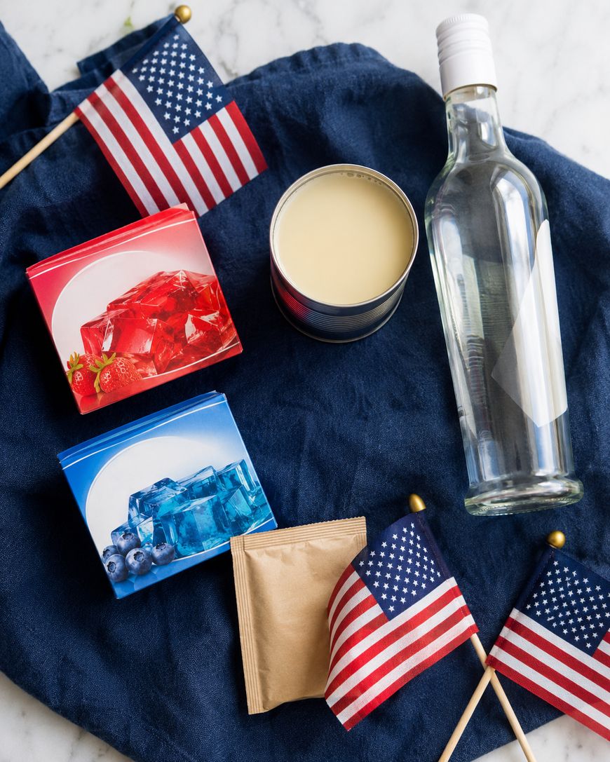 Two boxes of red and blue gelatin, a bottle of clear liquor, a small cup of white mixture, a brown packet, and three small American flags arranged on a dark blue cloth.