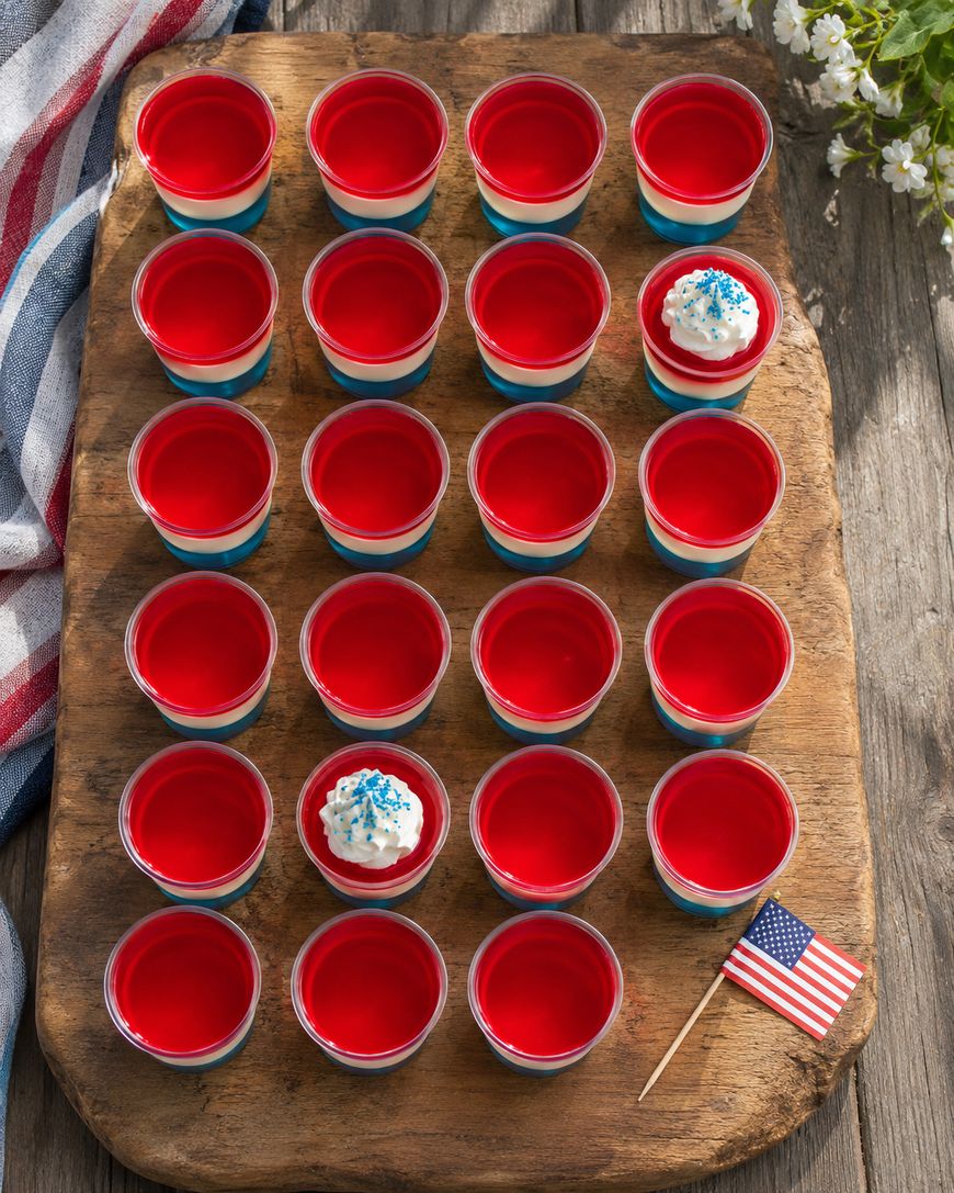 Small cups of layered red, white, and blue jello are arranged in a rectangle on a wooden tray. Two cups have whipped cream and blue sprinkles, and a small American flag is on the trays corner.