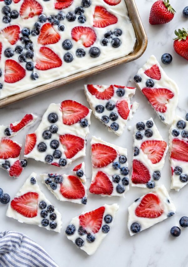 Yogurt bark topped with sliced strawberries and blueberries, broken into pieces on a marble surface. This red white and blue snack is perfect for patriotic snacks, with a baking tray and scattered fresh berries also visible.