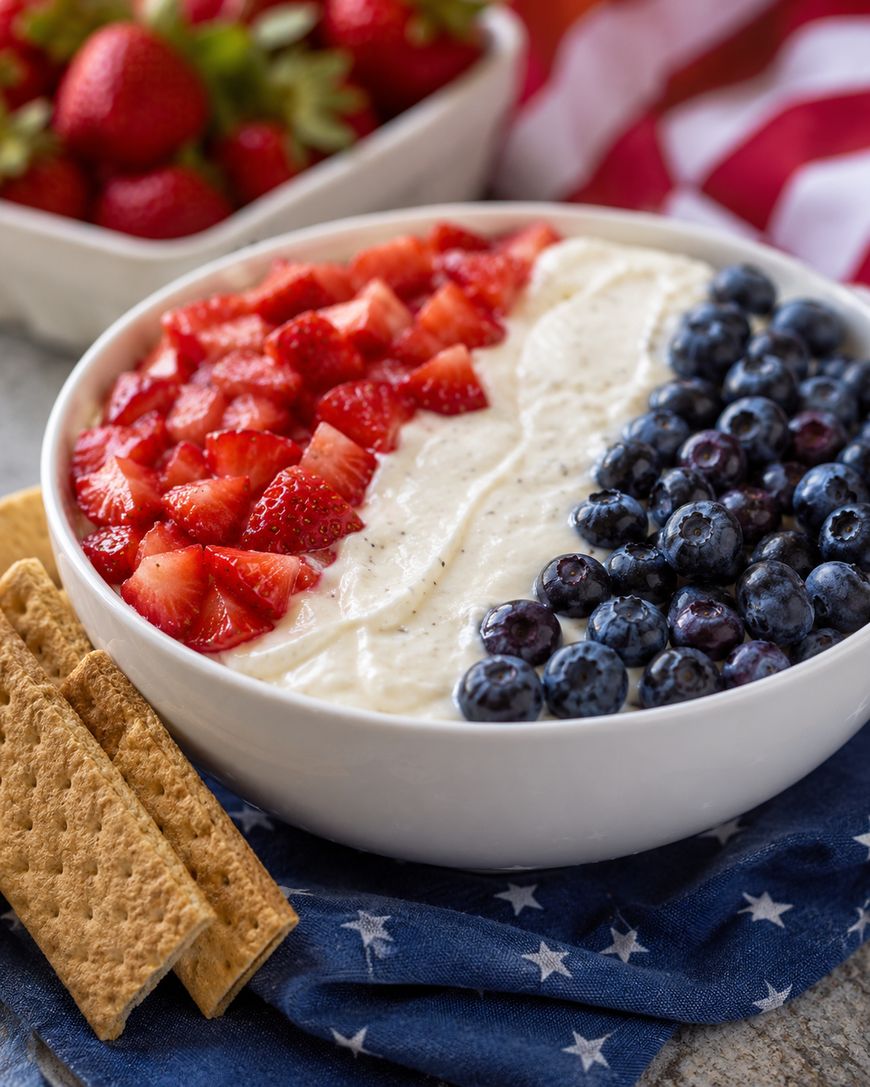 A bowl of creamy dip topped with chopped strawberries and blueberries arranged to resemble the American flag, with graham crackers on the side, placed on a blue star-patterned napkin.