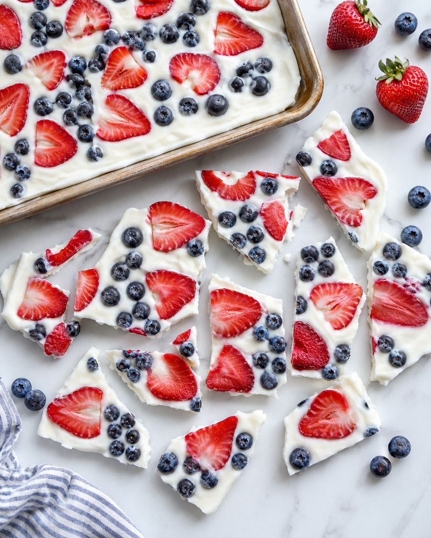 Broken pieces of frozen yogurt bark topped with sliced strawberries and blueberries are arranged on a marble surface next to a baking tray, with whole strawberries and blueberries scattered around.