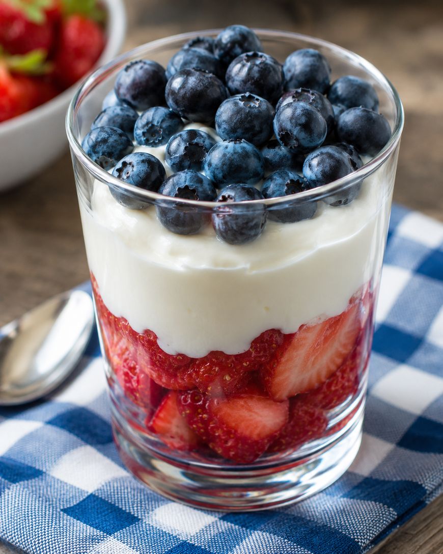 A glass filled with layers of sliced strawberries, creamy yogurt, and fresh blueberries, placed on a blue and white checkered napkin with a spoon nearby. A bowl of strawberries is in the background.