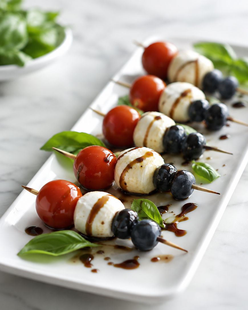 Skewers with cherry tomatoes, mozzarella balls, and blueberries on a white plate, garnished with fresh basil leaves and drizzled with balsamic glaze.