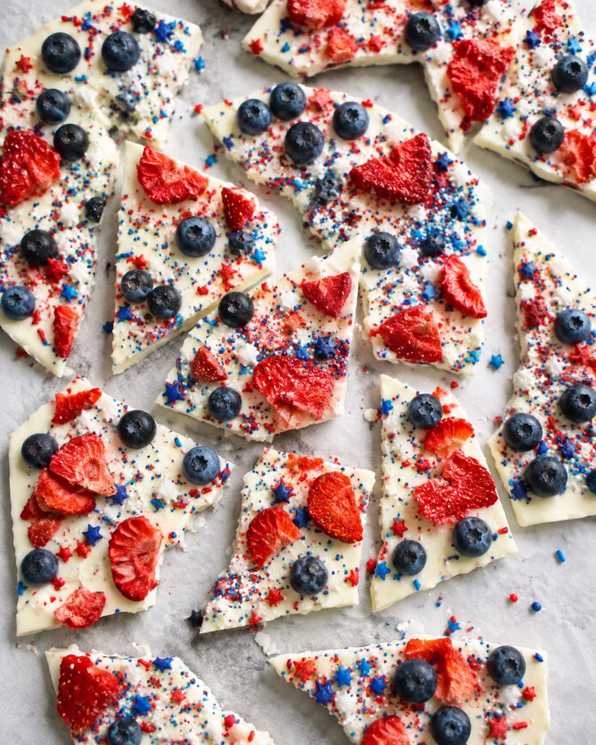 Triangular pieces of white chocolate bark topped with blueberries, dried strawberries, and red, white, and blue sprinkles, arranged on a light surface.