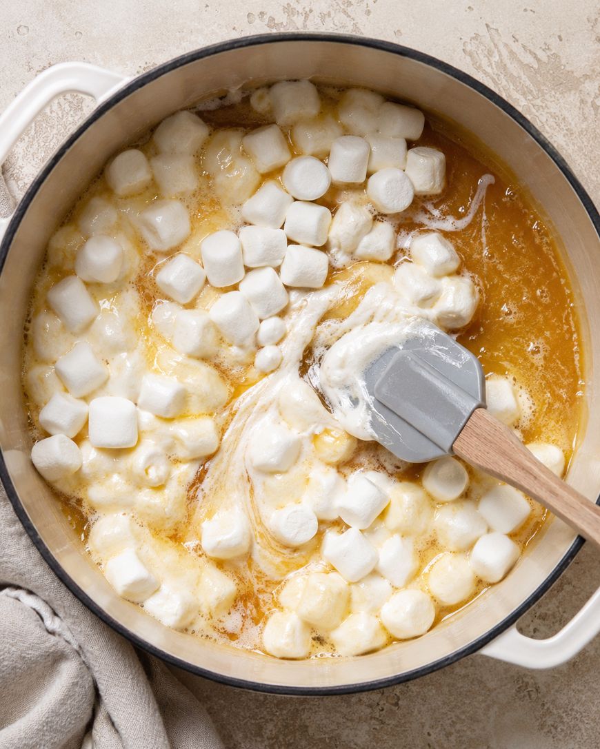 A pot with partially melted mini marshmallows being stirred into a golden liquid, likely melted butter, with a gray spatula on a light countertop and a beige towel beside it.