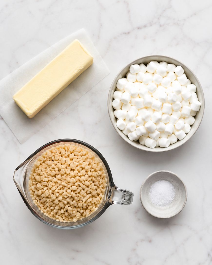 A stick of butter, a bowl of mini marshmallows, a measuring cup of crispy rice cereal, and a small dish of salt arranged on a white marble surface.
