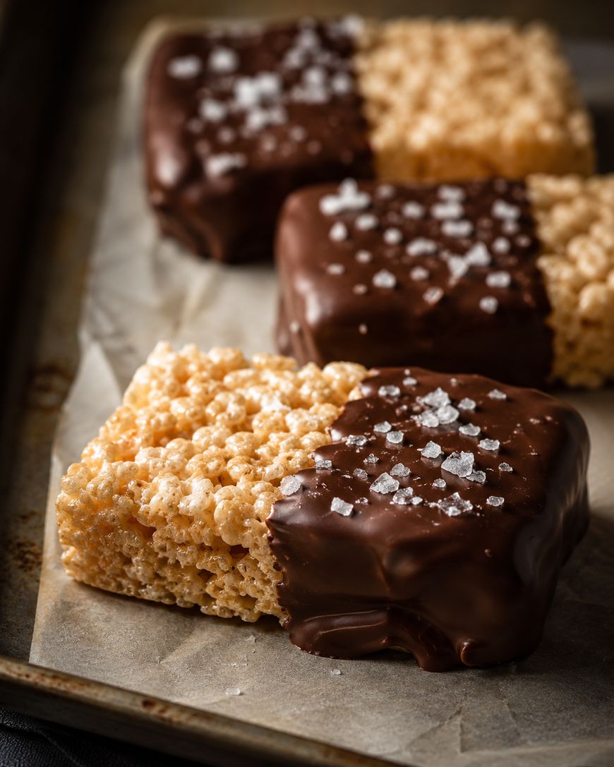 Three rectangular rice crispy treats, each half-dipped in chocolate and sprinkled with sea salt, are arranged in a row on parchment paper.