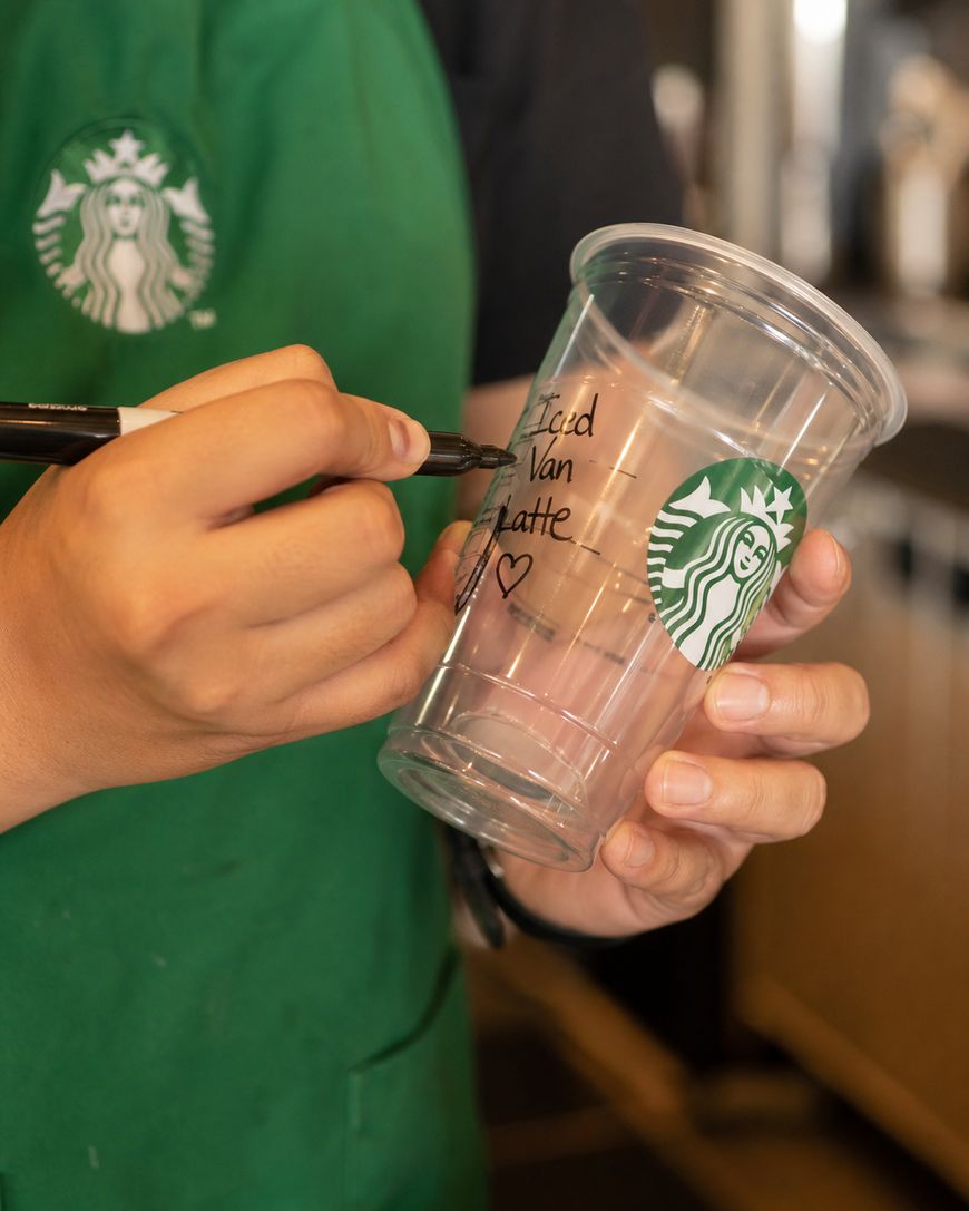 A person wearing a green Starbucks apron writes Iced Van Latte ♥ on a clear Starbucks cup with a black marker.