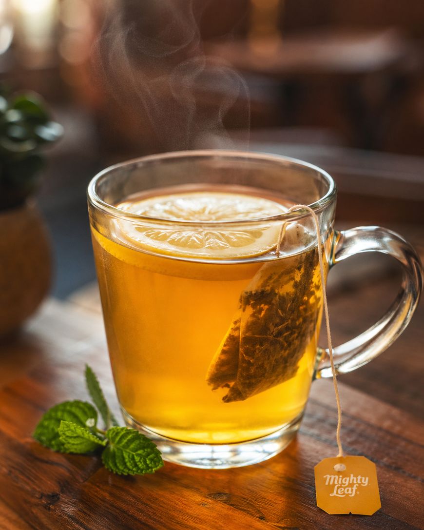A clear glass mug of hot tea with a tea bag labeled Mighty Leaf, garnished with a lemon slice. Steam rises from the tea. Fresh mint leaves rest on the wooden table beside the mug.