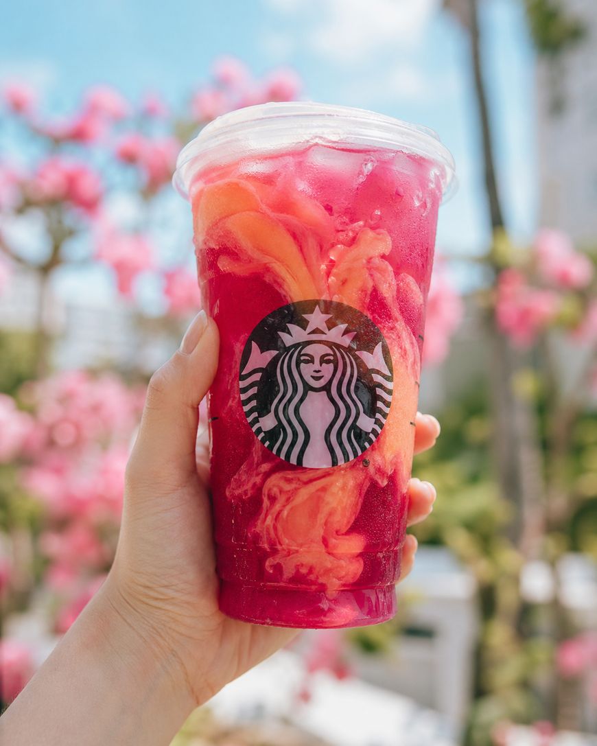 A hand holds a colorful Starbucks drink with vibrant pink and orange swirls in a clear cup, with blurred pink flowers and greenery in the sunny background.