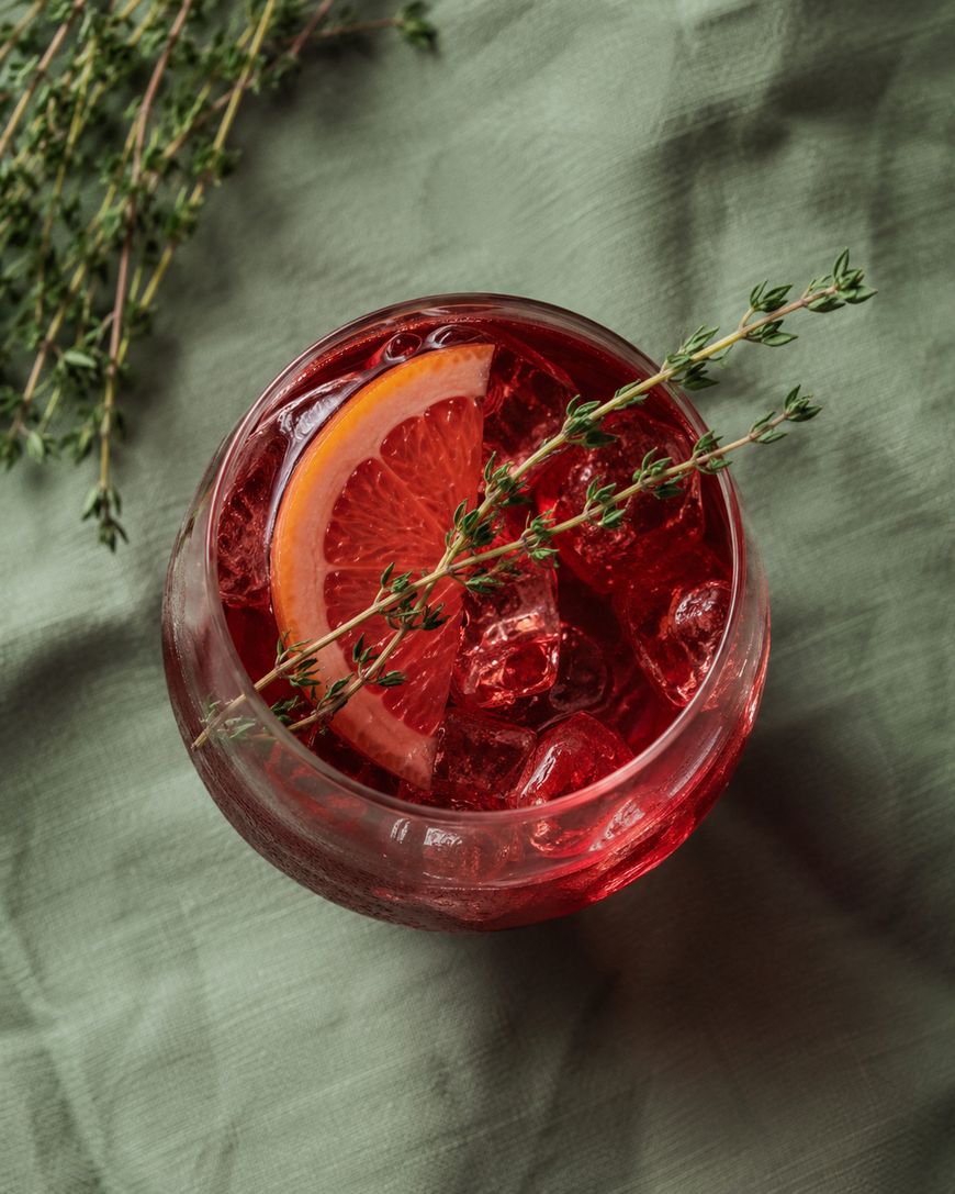 A glass of red iced drink garnished with a slice of orange and a sprig of thyme, placed on a green fabric surface with additional thyme sprigs nearby.