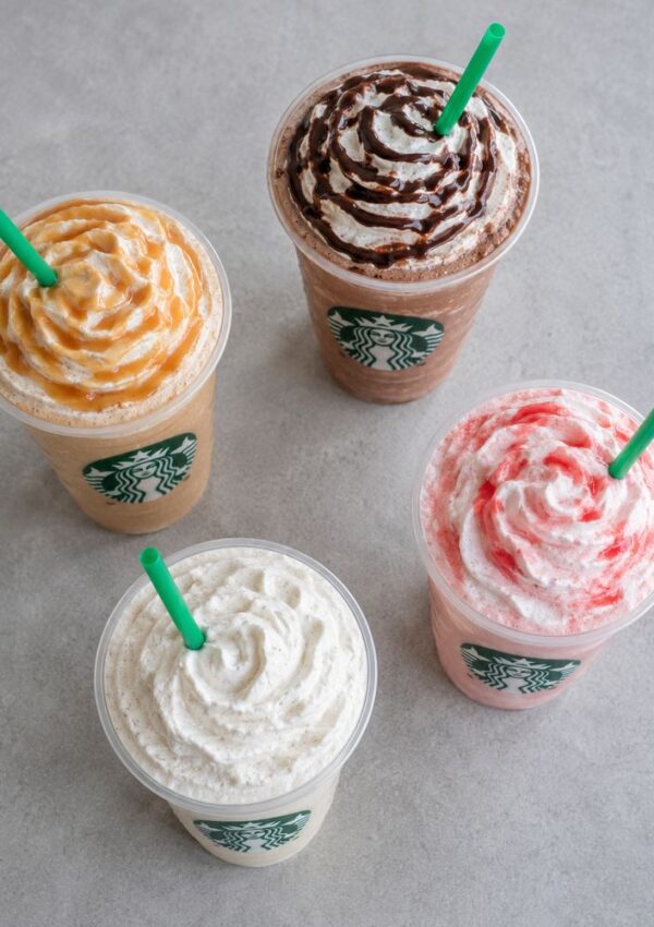 Four Starbucks Frappuccinos with whipped cream and colorful toppings—caramel, chocolate, strawberry, and plain—are arranged on a gray surface with green straws, showcasing some of the best Frappuccino orders among popular Starbucks drinks.