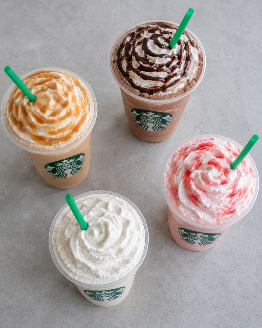 Four Starbucks frappuccinos on a gray surface, each topped with whipped cream and a different drizzle: caramel, chocolate, strawberry, and plain, with green straws in clear plastic cups.