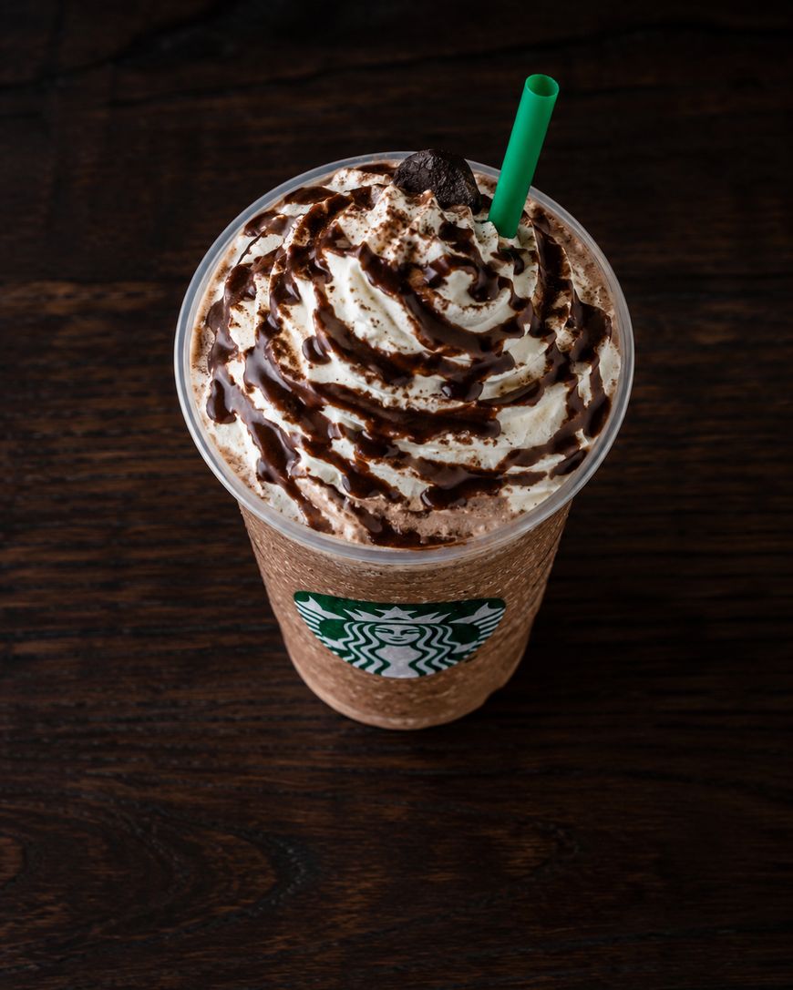 A Starbucks frappuccino topped with whipped cream, chocolate drizzle, and a green straw, shown on a dark wooden surface.