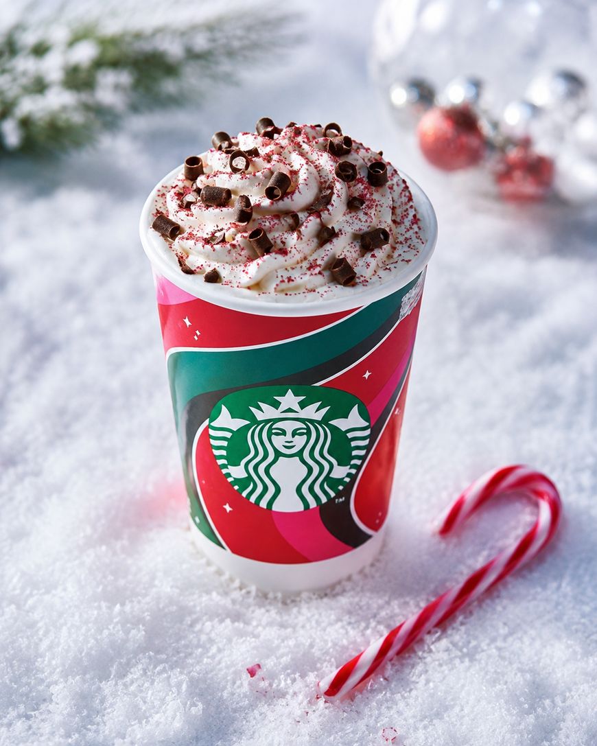 A festive Starbucks cup filled with whipped cream and chocolate curls sits on snow, with a candy cane in front and Christmas ornaments and pine branches blurred in the background.