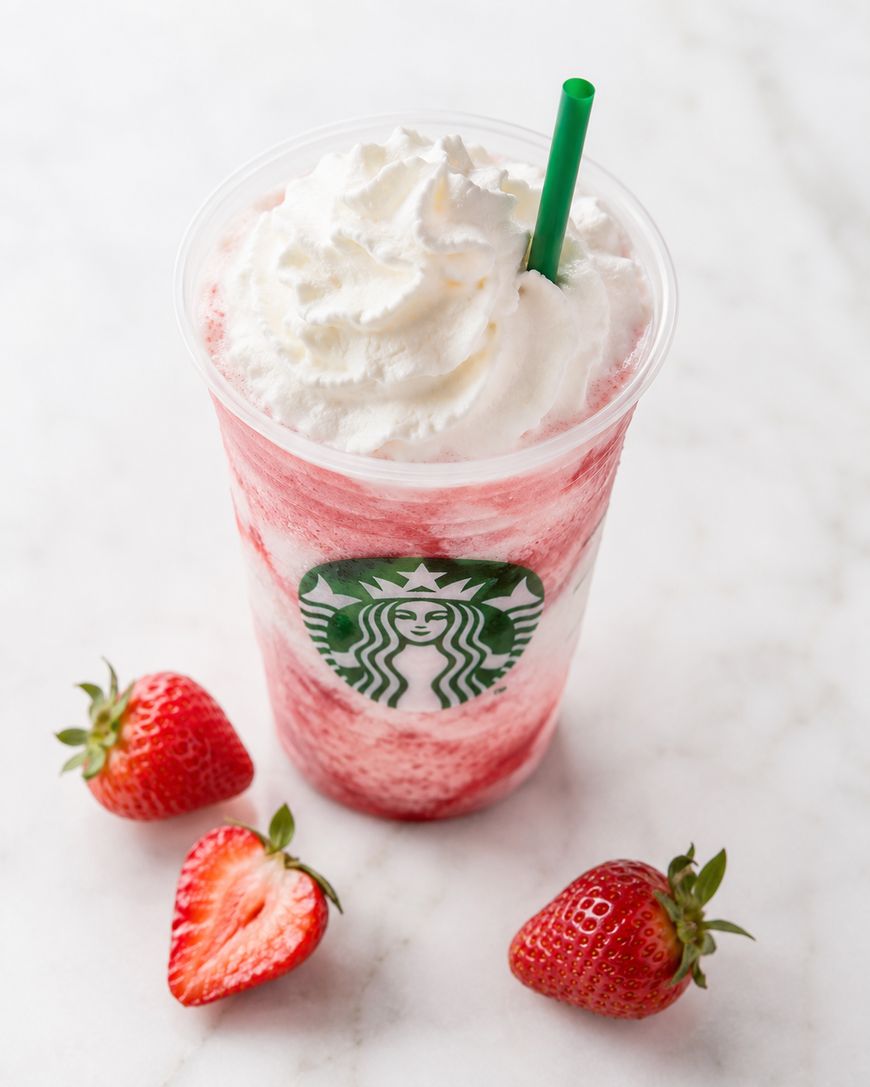 A Starbucks cup filled with a pink and white blended strawberry drink topped with whipped cream, with a green straw. Three fresh strawberries, one sliced in half, are placed beside the cup on a white marble surface.