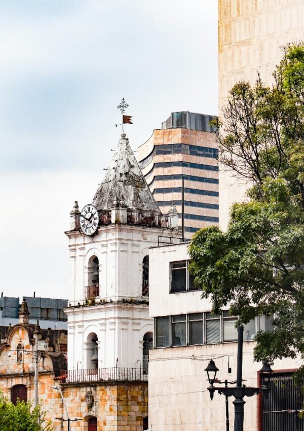 A white clock tower with a weather vane rises among modern and historic buildings, partially framed by green trees under a cloudy sky.