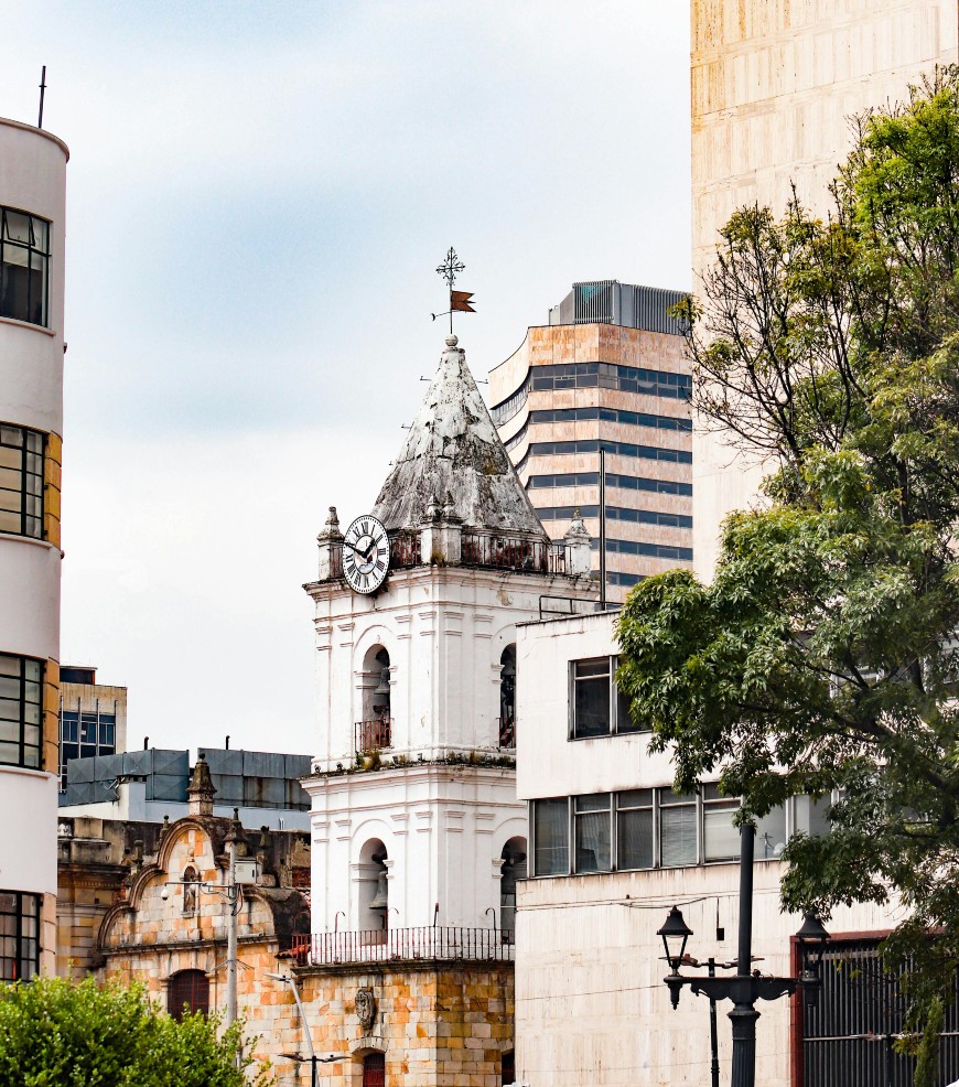 A white clock tower with a pointed roof stands among modern and older buildings, with green trees in the foreground and a partly cloudy sky above.