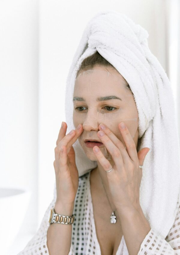 A woman with a towel wrapped around her head applies a sheet face mask, embracing summer beauty trends while wearing a white robe and jewelry, standing in a bright, minimalist bathroom.