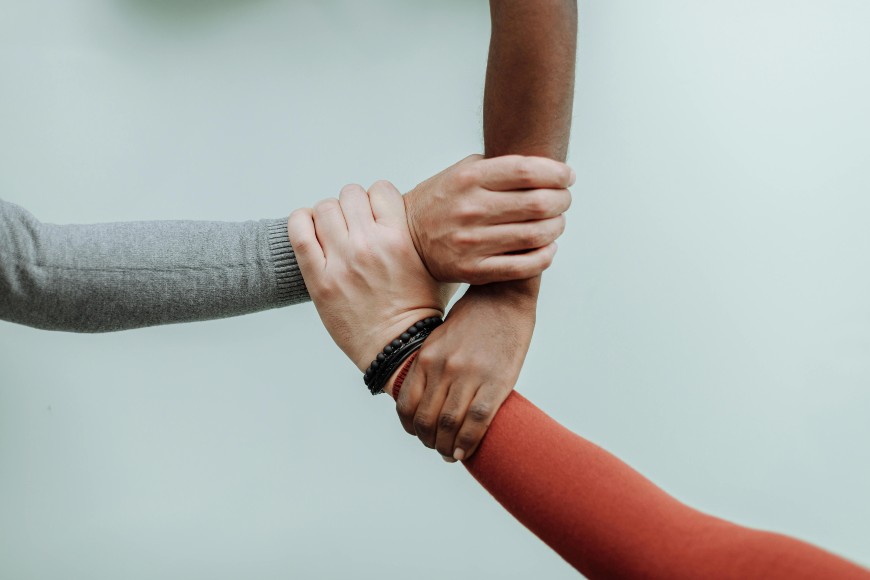 Three arms of different skin tones are interlocked, with each hand grasping anothers wrist against a plain light background, symbolizing unity and teamwork.