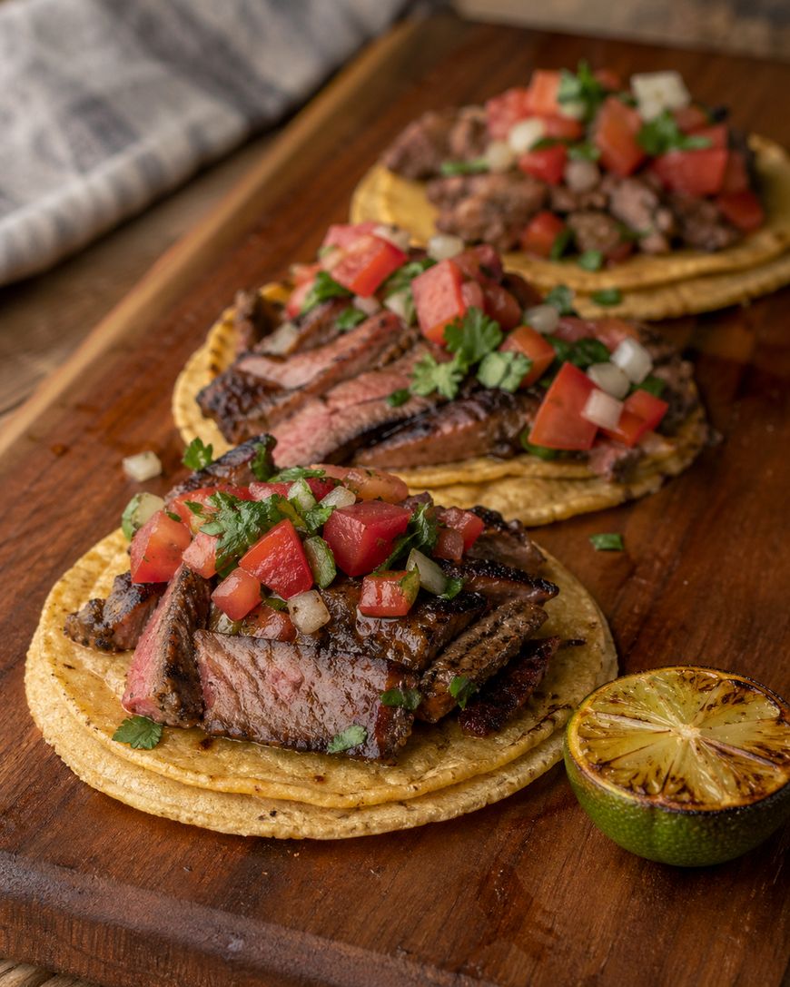 Three steak tacos topped with diced tomatoes, onions, and cilantro sit on a wooden board, next to a grilled lime half. A striped cloth is in the background.