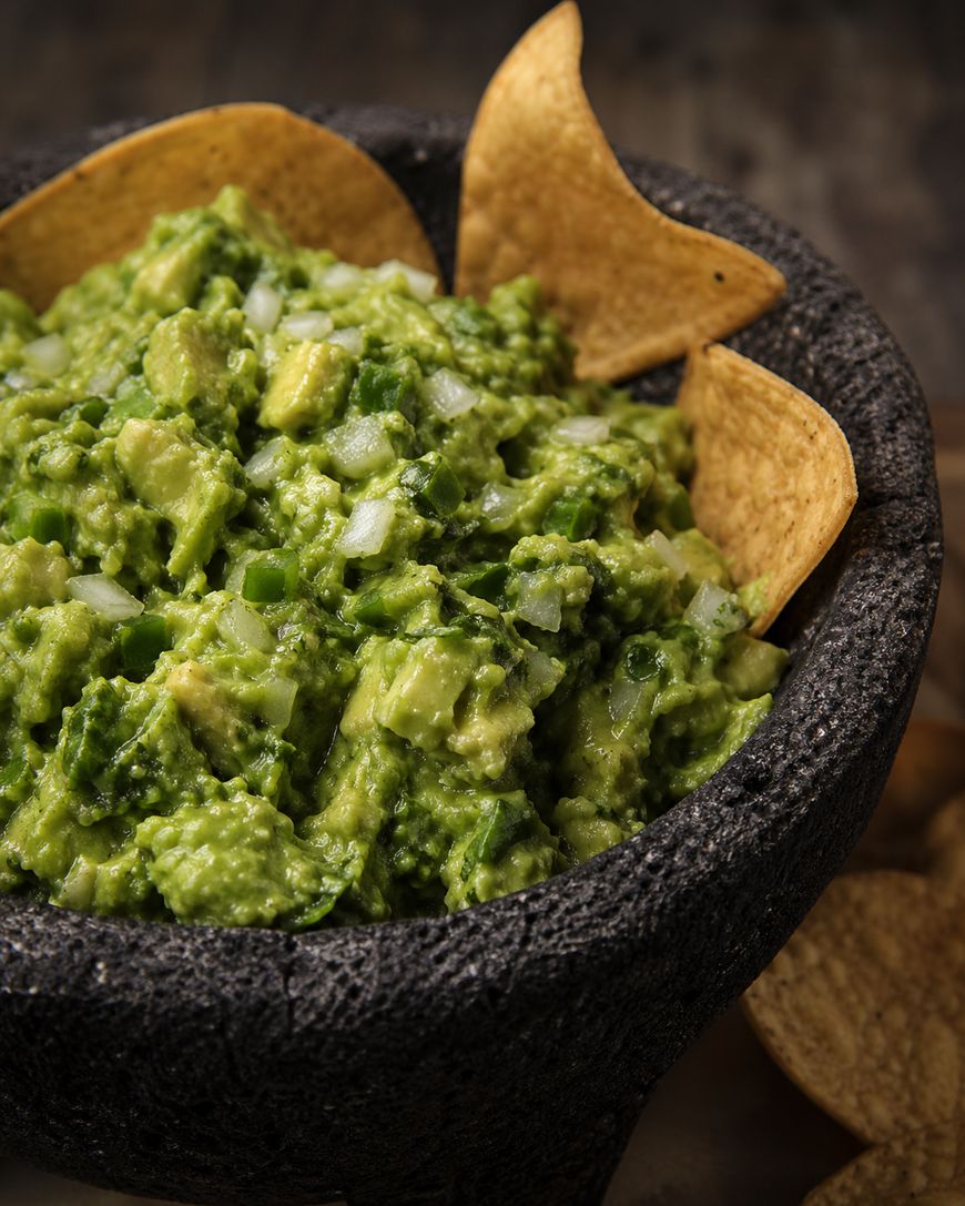 A stone bowl filled with chunky guacamole garnished with diced onions and surrounded by tortilla chips.