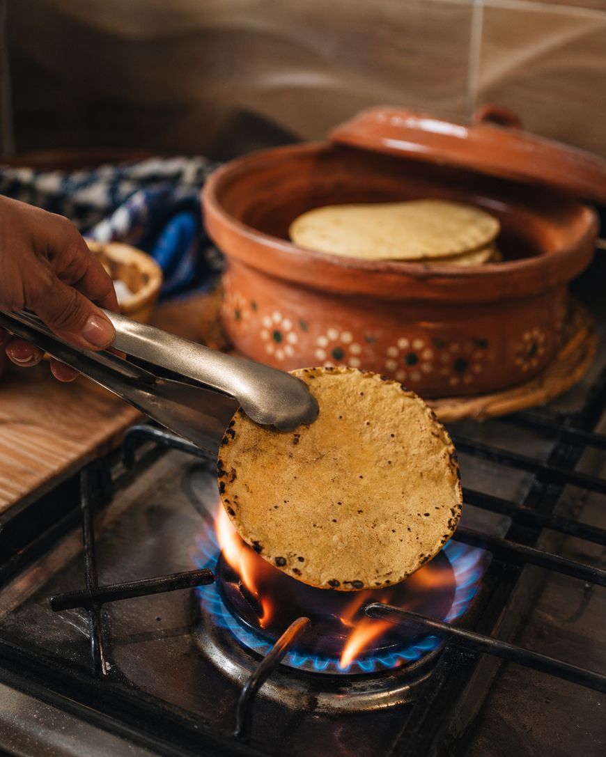 A hand holds tongs to heat a corn tortilla over a gas stove flame. In the background, a clay pot with more tortillas sits on the counter.