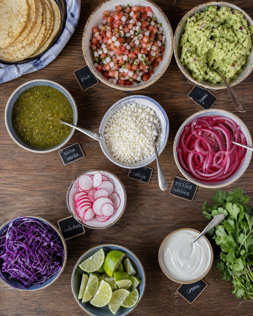 A top view of various taco toppings in bowls, including pico de gallo, guacamole, salsa verde, cotija cheese, pickled onions, sliced radishes, shredded purple cabbage, lime wedges, crema, cilantro, and corn tortillas.
