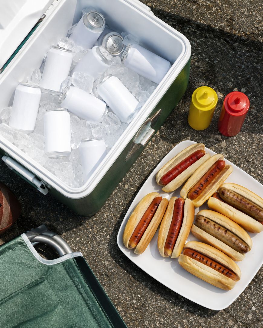 A cooler filled with ice and canned drinks sits next to a plate of grilled hot dogs in buns, with ketchup and mustard bottles nearby on a concrete surface. A green chair is visible in the corner.