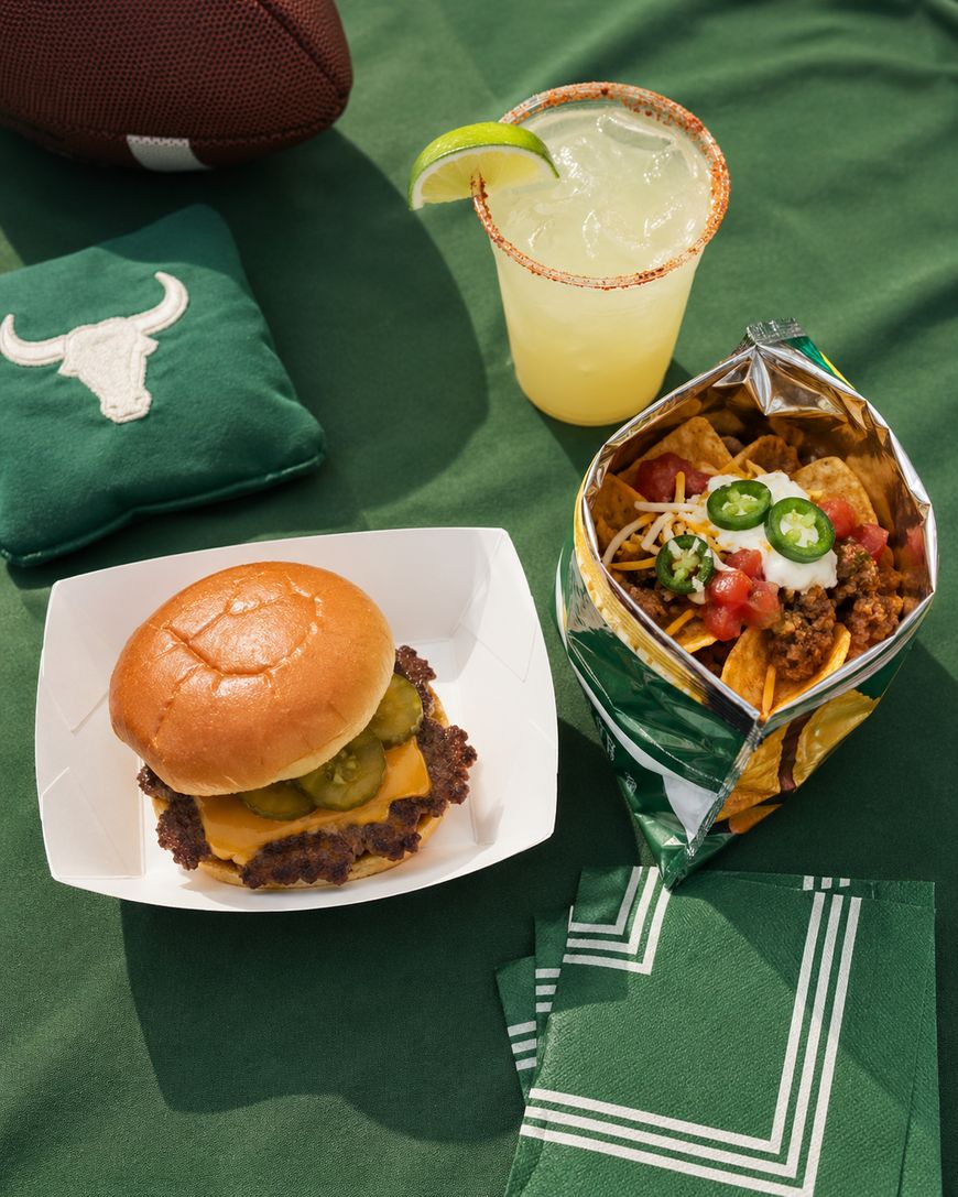 A cheeseburger with pickles, a margarita with a lime wedge, and a bag of nachos topped with ground beef, diced tomato, onions, jalapeños, and cheese, set on a green table with football-themed napkins and a football.